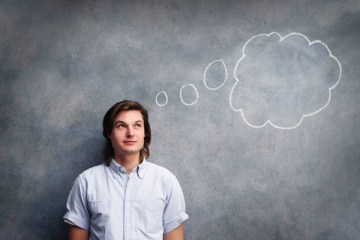 A young man stands in front of a chalk board with a thought bubble above his head.